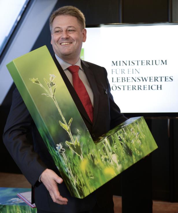 Ein lächelnder Mann im Anzug hält eine grüne, L-förmige Box mit Pflanzenmotiven.