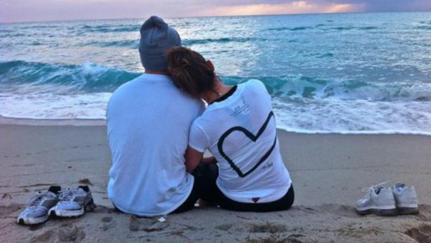Ein Paar sitzt am Strand und blickt auf das Meer. Ihre Schuhe stehen im Sand.