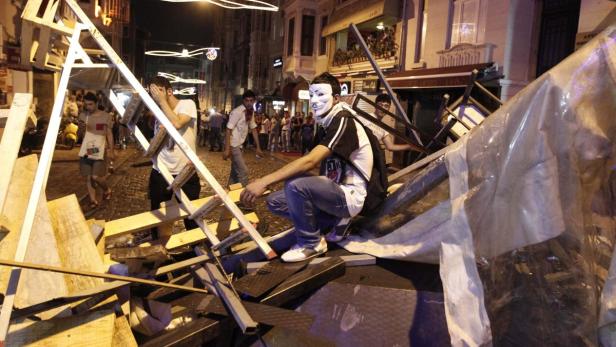 Ein Demonstrant mit einer Maske sitzt auf einer Barrikade in einer belebten Straße.
