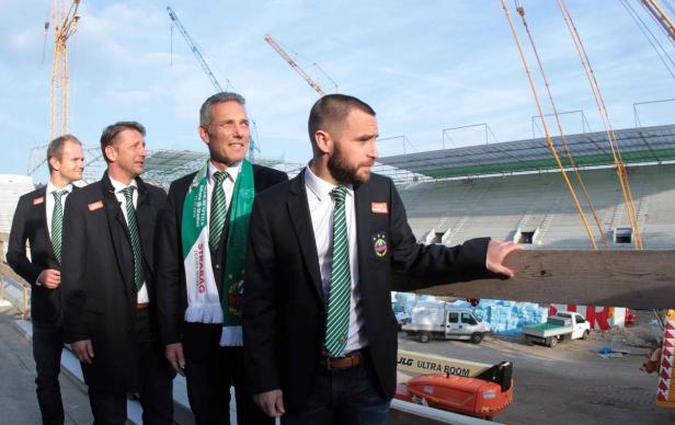 Eine Gruppe von Männern in Anzügen steht vor einem im Bau befindlichen Stadion.