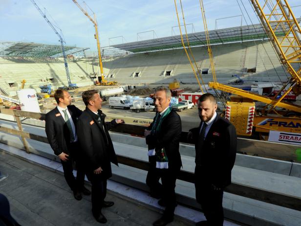 Vier Männer in Anzügen vor einer im Bau befindlichen Sportarena mit Kränen.