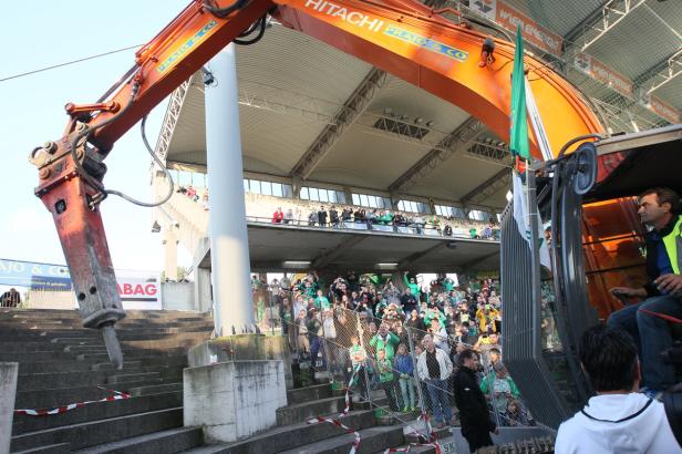 Ein Bagger mit Abbruchhammer vor einer Zuschauertribüne voller Menschen.