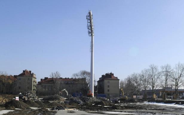 Ein hoher Sendemast steht auf einem Gelände mit Baumaschinen und Wohnhäusern im Hintergrund.