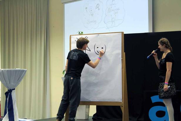 Ein Mann zeichnet ein Gesicht auf ein Whiteboard, während eine Frau mit einem Mikrofon daneben steht.
