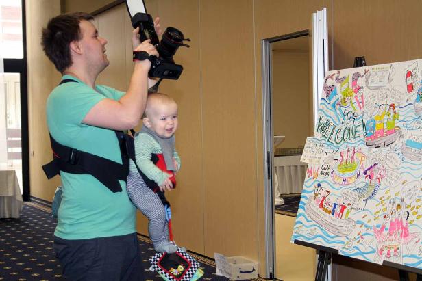 Ein Mann fotografiert mit einer Kamera, während er ein Baby in einer Trage trägt.