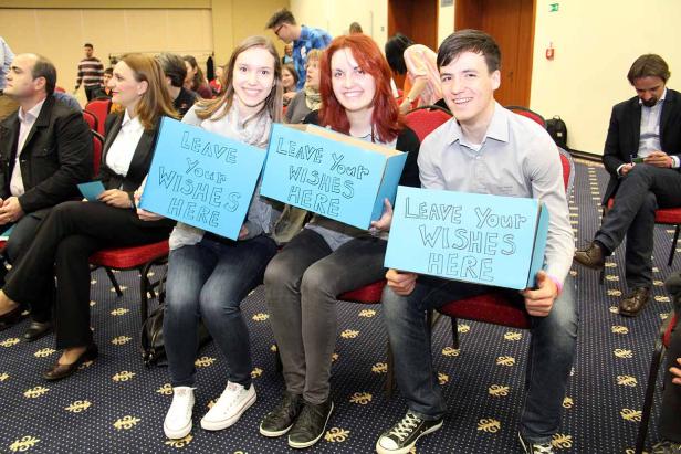 Drei junge Leute sitzen in einem Raum und halten blaue Schilder mit der Aufschrift „Leave Your Wishes Here“.