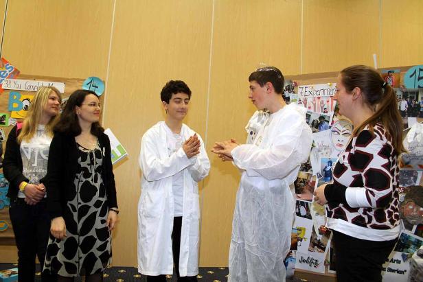 Zwei Jungen in Laborkitteln stehen vor einer Wand mit Postern, umgeben von drei Frauen.