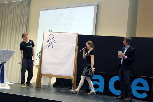 Eine Frau spricht auf einer Bühne, während ein Mann ein Strichmännchen auf ein Whiteboard zeichnet.