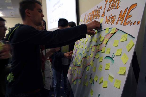 Eine Gruppe von Menschen klebt Notizen auf ein großes Poster mit der Aufschrift „A Learning I Take With Me…“.