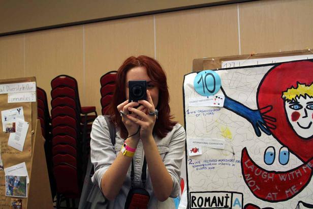 Eine Frau mit rotem Haar fotografiert mit einer Kompaktkamera vor einem Poster über Studentenprojekte.