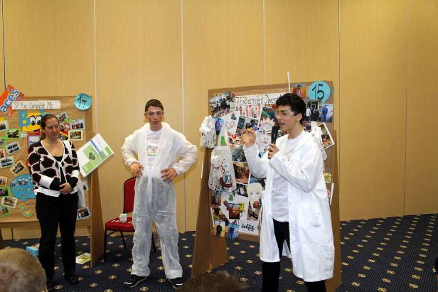 Zwei Jugendliche in Laborkitteln präsentieren ein Projekt vor einer Frau.