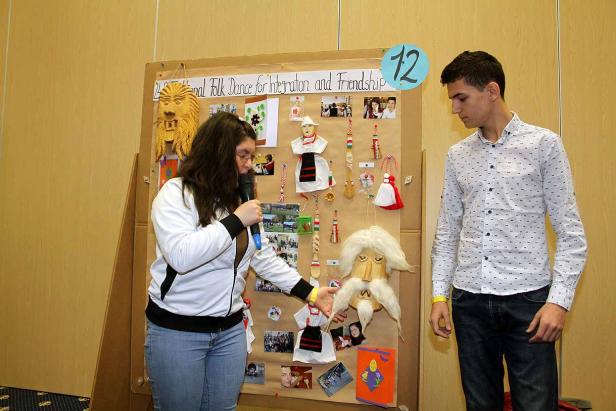 Eine junge Frau präsentiert mit Mikrofon ein Projekt über Volkstänze und Freundschaft vor einem jungen Mann.