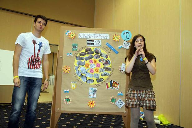 Eine junge Frau spricht vor einem Publikum, neben ihr steht ein junger Mann und eine Präsentationstafel.