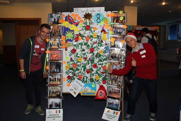 Zwei Personen posieren vor einer Tafel mit dem Motto „Diversity makes rich“ und Fotos zum Thema Vielfalt.