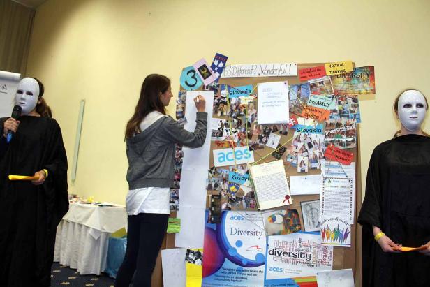 Eine Frau schreibt an einer Pinnwand, die mit Fotos und dem Wort „Diversity“ geschmückt ist. Zwei Personen mit weißen Masken stehen daneben.