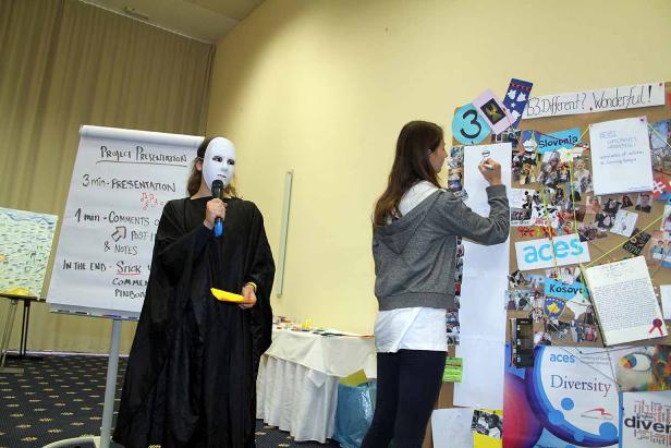 Eine Person mit Maske hält ein Mikrofon, während eine andere an einer Pinnwand arbeitet.