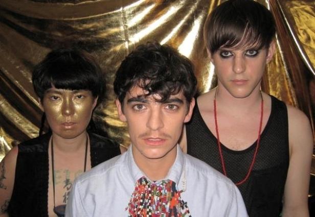 Drei Personen posieren vor einem goldenen Hintergrund, eine mit goldener Farbe im Gesicht.