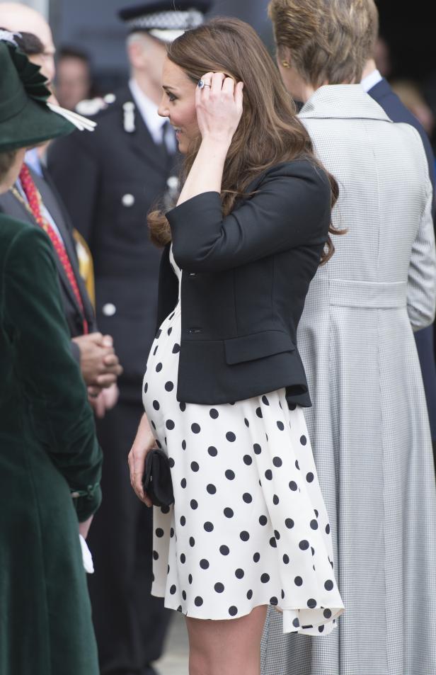 Eine schwangere Frau in einem gepunkteten Kleid und Blazer steht im Freien.