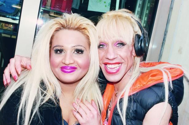 Zwei Personen mit blonden Haaren und auffälligem Make-up posieren für ein Foto.