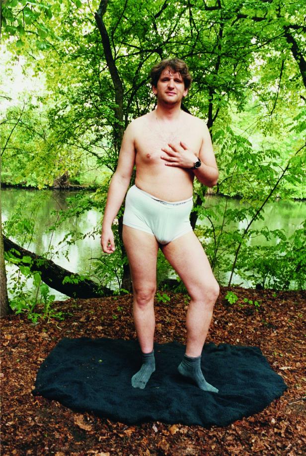 Ein Mann posiert in Unterwäsche und Socken vor einem Wald und einem See.
