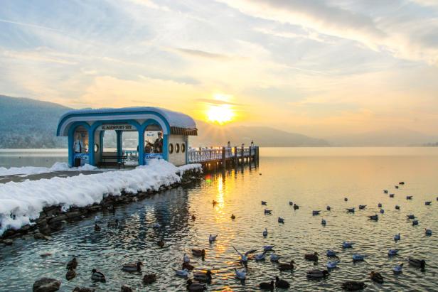 Ein schneebedeckter Steg am Klagenfurt See bei Sonnenaufgang, mit vielen Vögeln im Wasser.