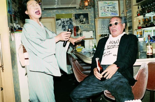 Eine Frau im Kimono singt in einem Lokal, während ein Mann mit Sonnenbrille und Bier zuhört.