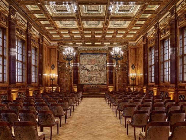 Ein holzgetäfelter Saal mit Stuhlreihen und einem großen Wandteppich.