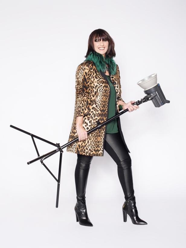 Eine Frau im Leopardenmantel hält eine Studioleuchte und ein Stativ.