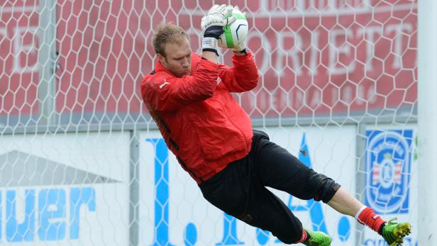 Ein Torwart in roter Jacke fängt einen Ball vor einem Fußballtor.