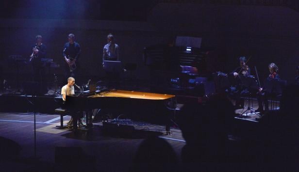 Ein Mann spielt Klavier und singt auf einer Bühne mit anderen Musikern.