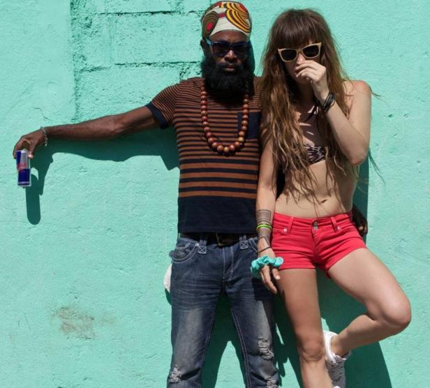 Ein Mann mit Turban und Bart und eine Frau mit roter Hose posieren vor einer türkisfarbenen Wand.