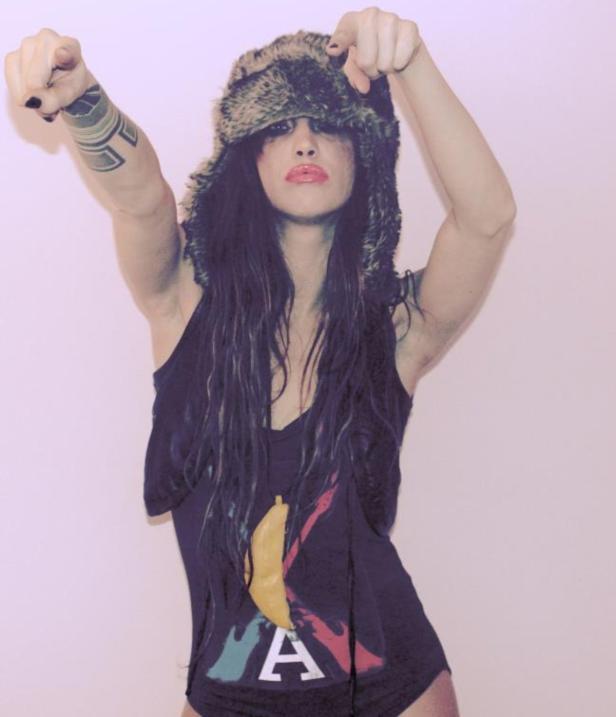 Eine Frau mit Fellmütze und Tattoo auf dem Arm zeigt mit beiden Händen auf den Betrachter.