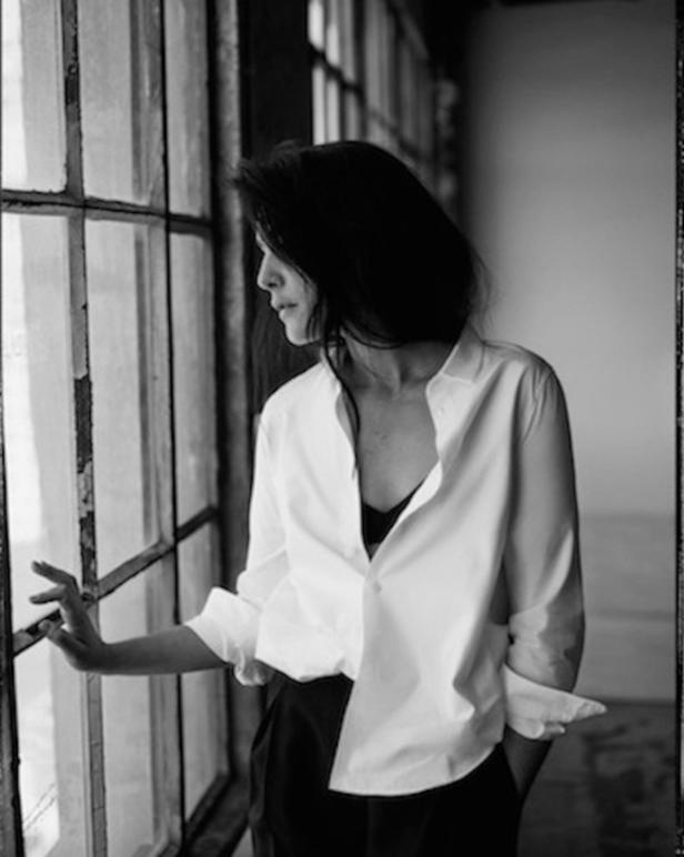 Eine Frau in Hemd und Hose steht am Fenster und schaut nach draußen.