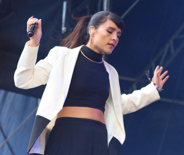 Eine Sängerin tritt in einem weißen Blazer und schwarzem Top auf einer Bühne auf.