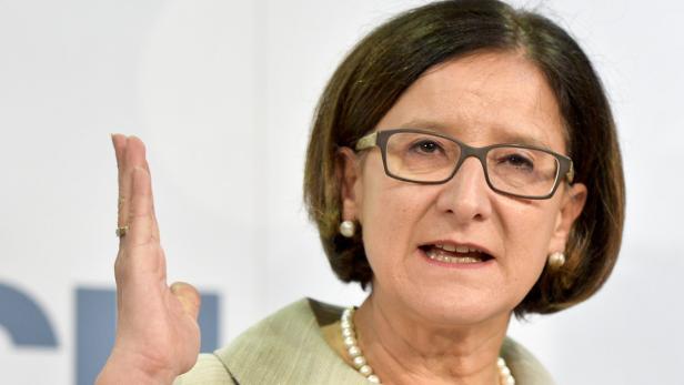 Eine Frau mit Brille und Perlenkette hebt ihre Hand.