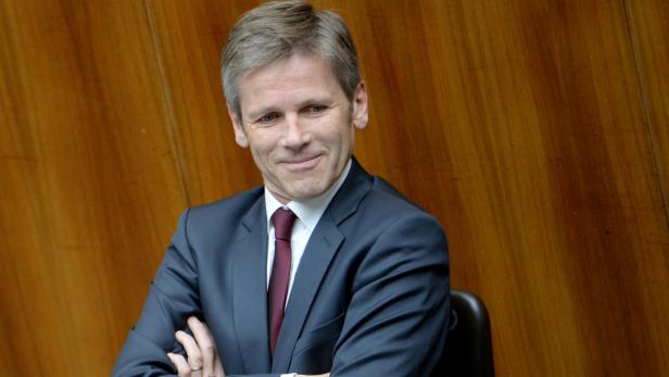 Ein Mann im Anzug sitzt vor einer hölzernen Wand.