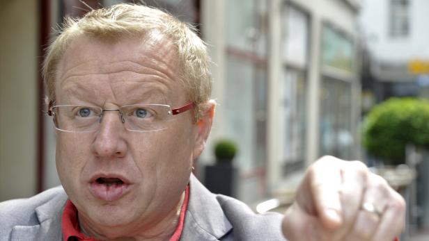 Ein Mann mit Brille gestikuliert vor einem unscharfen Hintergrund.