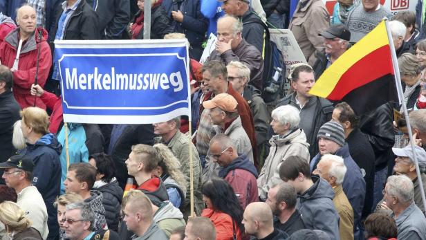 Eine Menschenmenge demonstriert mit einer Deutschlandflagge und einem Schild mit der Aufschrift „Merkel muss weg“.