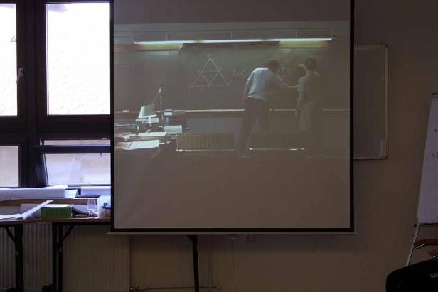 Eine Projektion zeigt zwei Personen, die an einer Tafel mit geometrischen Figuren arbeiten.