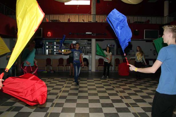 Mehrere Personen schwingen bunte Tücher in einem Raum mit Schachbrettmusterboden.