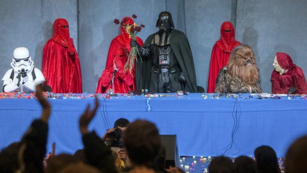 Eine Gruppe von Personen, verkleidet als Star Wars-Charaktere, sitzt an einem Tisch vor einer Menschenmenge.