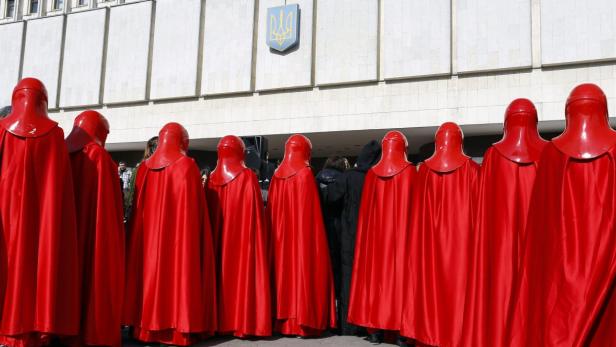 Eine Gruppe von Personen in roten Kostümen steht vor einem Gebäude mit dem ukrainischen Wappen.