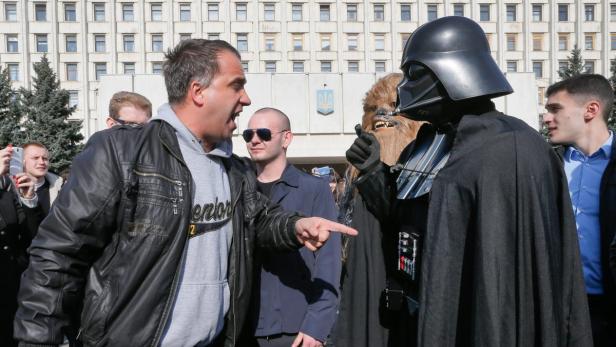 Ein Mann gestikuliert vor Darth Vader und Chewbacca vor einem Gebäude.