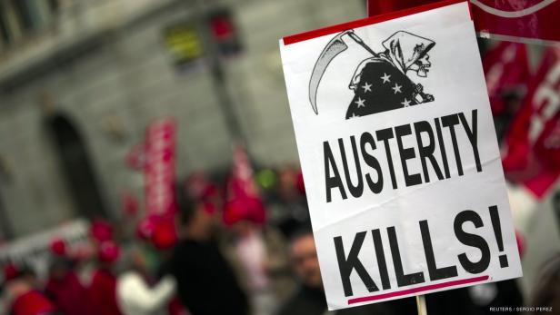 Ein Schild mit der Aufschrift „Austerity Kills!“ und einer Zeichnung des Sensenmanns wird auf einer Demonstration gezeigt.