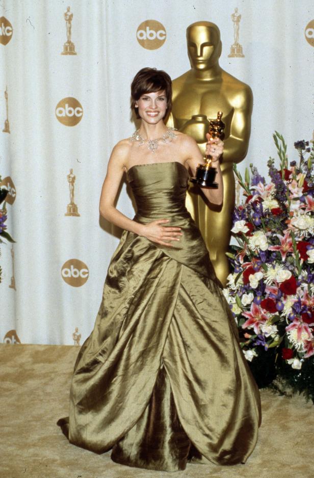 Hilary Swank mit einem Oscar in der Hand vor einer goldenen Statue.