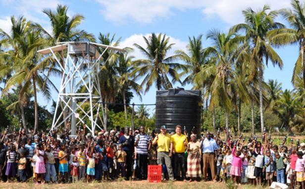 Eine große Gruppe Menschen steht vor einem Wasserturm und einem Wassertank unter Palmen.