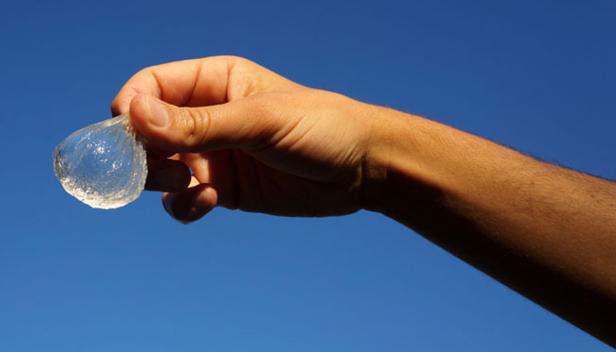 Eine Hand hält eine durchsichtige Wasserbombe vor einem blauen Himmel.