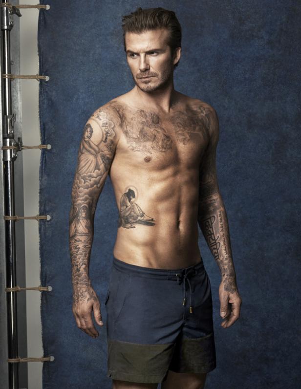 David Beckham posiert mit freiem Oberkörper und zahlreichen Tätowierungen vor einem blauen Hintergrund.