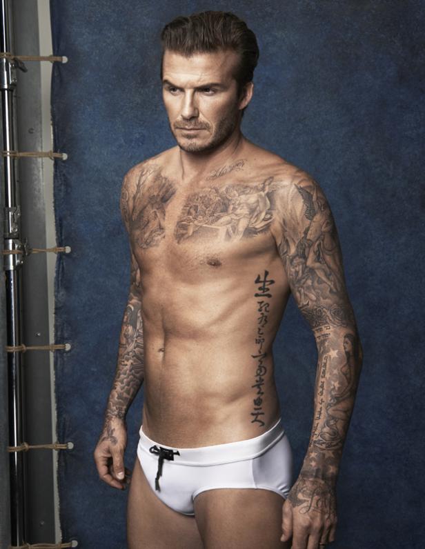 David Beckham mit zahlreichen Tätowierungen und Badehose vor blauem Hintergrund.