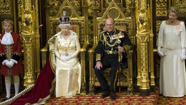 Königin Elisabeth II. und Prinz Philip sitzen auf goldenen Thronen, flankiert von zwei Begleitern.